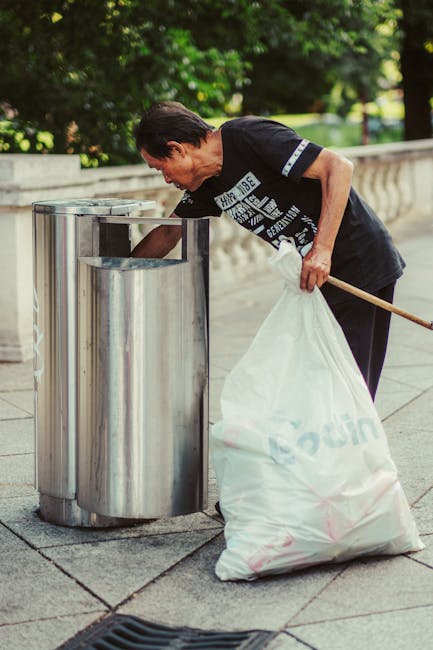 A man with short dark hair, wearing a black T-shirt with white text and dark trousers, is leaning over a public metal waste receptacle on a paved sidewalk, actively disposing of rubbish. He is holding a large white plastic bag filled with waste in one hand and using the other hand to reach into the recycling bin. The metal bins are cylindrical with a shiny, reflective surface and are positioned side by side, set against a backdrop of greenery, including leafy trees and bushes, indicating an outdoor urban environment. The scene is well-lit with natural daylight, highlighting the textures of the concrete pavement, the metallic finish of the bins, and the casual attire of the individual. This image depicts onsite rubbish collection and disposal, related to private waste management services, such as those offered by Rubbish Clearance Belgravia, supporting environmentally responsible waste handling outside of traditional municipal collection methods.