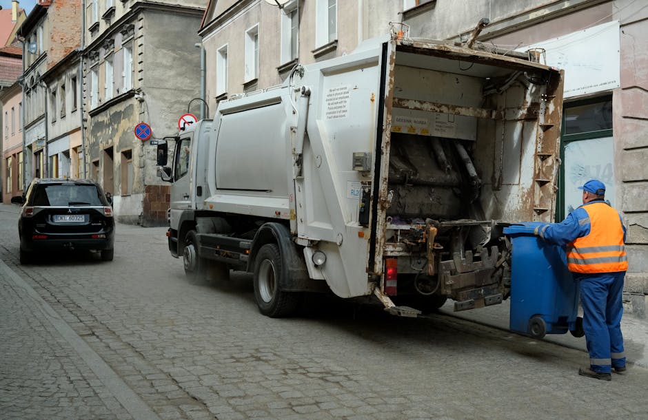 A white waste collection truck with rust and wear visible on its rear compactor section is parked on a narrow cobblestone street, situated alongside old, multi-story buildings with a weathered facade. The truck's rear is open, revealing mechanical components used for compacting and lifting rubbish. A worker dressed in blue overalls, a bright orange high-visibility vest, and a blue cap is positioned on the right side, actively pushing a mobile blue waste bin towards the truck's open compactor area. In the background, a black car is parked on the street, and above, there are several traffic signs mounted on the building's wall, indicating parking restrictions. The environment appears to be an urban area with a mix of historic architecture and modern waste collection services, reflecting an alternative, private approach to rubbish removal that complements typical municipal rubbish collection methods. Rubbish Clearance Belgravia may carry out similar on-site clearance or private waste handling for local shops and residents in Belgravia.