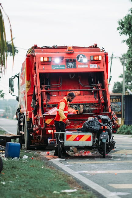 A sanitation worker wearing a red and yellow high-visibility vest is positioned next to the open back of a large red waste collection truck, which is parked on a street. The worker is engaged in emptying or sorting waste, with a black plastic bag placed nearby on the pavement. The truck's rear compartment is open, revealing mechanical components and a partially visible waste hopper. Surrounding the scene are elements typical of an urban environment, including a motorcycle parked close to the truck, a small wooden barrier with reflective red and yellow markings, and some scattered rubbish on the ground. In the background, there are trees, power lines, and a building that appears to be a small commercial establishment. The scene is illuminated by natural daylight, indicating an outdoor setting during daytime. This scene illustrates waste collection and rubbish removal activities, consistent with private or independent collection services such as those offered by Rubbish Clearance Belgravia.