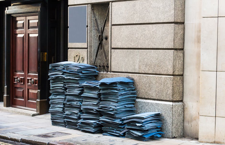 A large stack of blue paper files and documents is piled against the exterior stone wall of a building on a city sidewalk. The documents are stacked haphazardly, with some leaning slightly to one side, and appear to be fresh or recently gathered, exhibiting a slight sheen from the paper material. The wall behind the stacks is composed of rough-textured, grey concrete or stone blocks, with visible horizontal joints, and a metal security shutter with a traditional cross-shaped lock mechanism is partially visible behind the papers. To the left, there is a dark wooden double door with raised rectangular panels and metallic door knockers, set within a black frame. The ground is paved with square stone tiles, showing some weathering and minor cracks, typical of an urban environment. The lighting is natural, casting subtle shadows, indicating daylight conditions. This scene possibly indicates a private property handling bulk waste or document clearance, aligning with independent or alternative waste disposal practices often handled by professional rubbish removal services like Rubbish Clearance Belgravia.