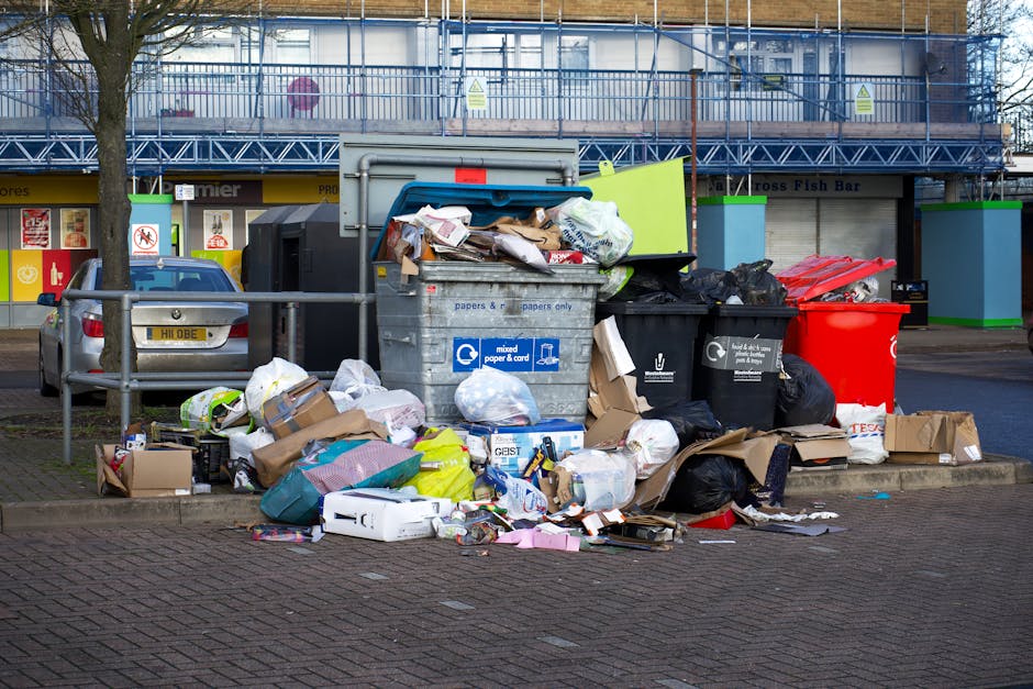 A large collection of overflowing rubbish bins and scattered trash bags situated on a paved urban street corner, with various types of waste including brown cardboard boxes, plastic bags, paper, and packaging materials spilling onto the ground. The recycling bins, mostly black with one red and one grey with blue label, are filled to capacity, causing debris to spill onto the surrounding pavement. Behind the rubbish area, there is a metal railing, and in the background, a row of shopfronts with signage and a building with gender-specific information signs and windows covered by blue safety netting. A parked silver car is visible to the left of the rubbish, and a leafless tree stands nearby. The scene is illuminated by natural daylight, highlighting the mess and the cluttered environment, which pertains to an area where private rubbish collection or on-site clearance may be required, reflective of the waste management context for Belgravia residents.